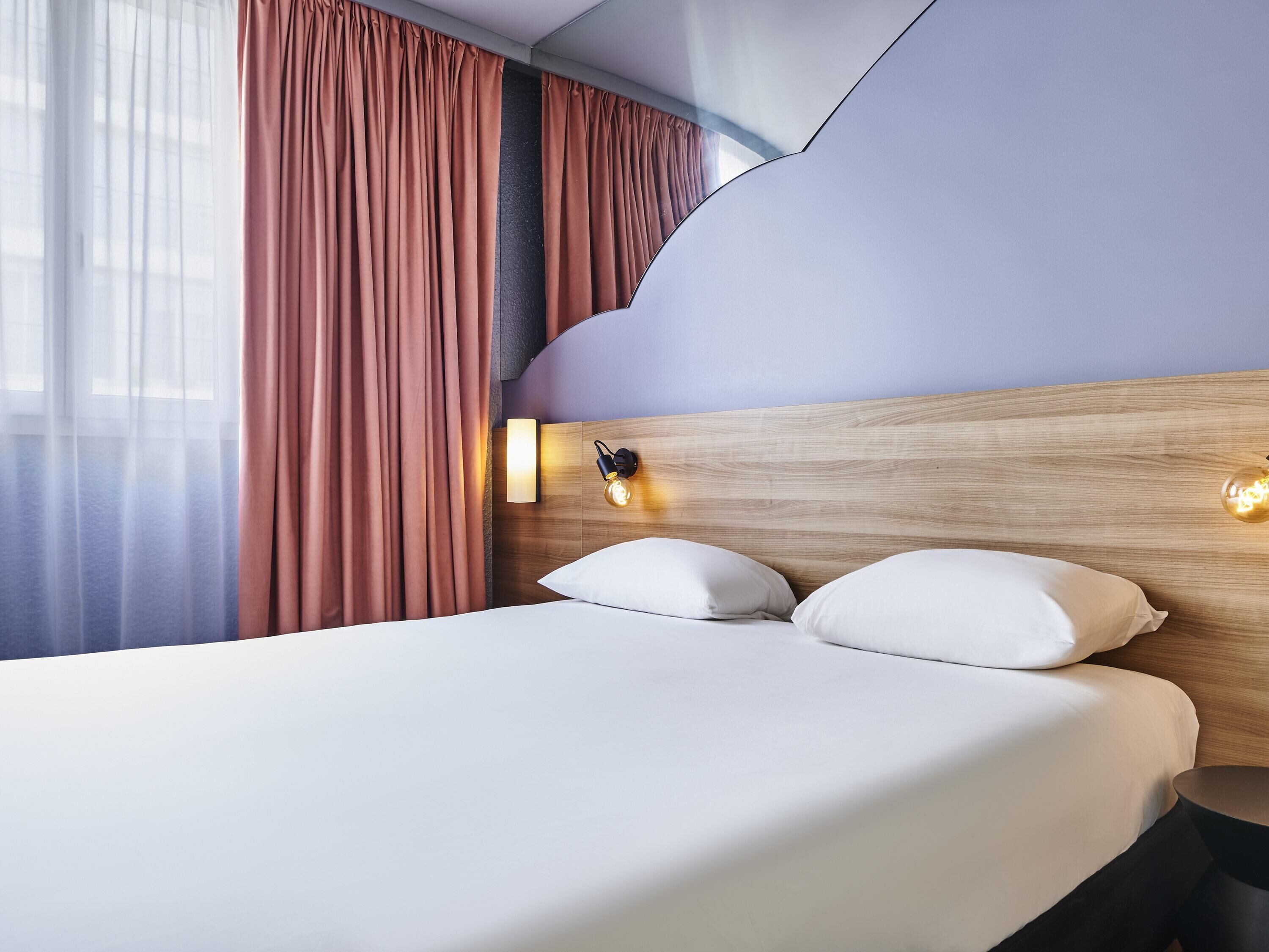 Hotellihuone, jossa on sänky, verhot, seinään kiinnitetty valaisin ja puinen pääty.
