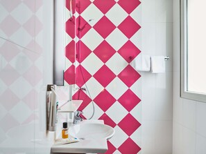 Une salle de bain avec un lavabo blanc, un miroir et une douche dont le mur est décoré d’un motif losangique rose et blanc.