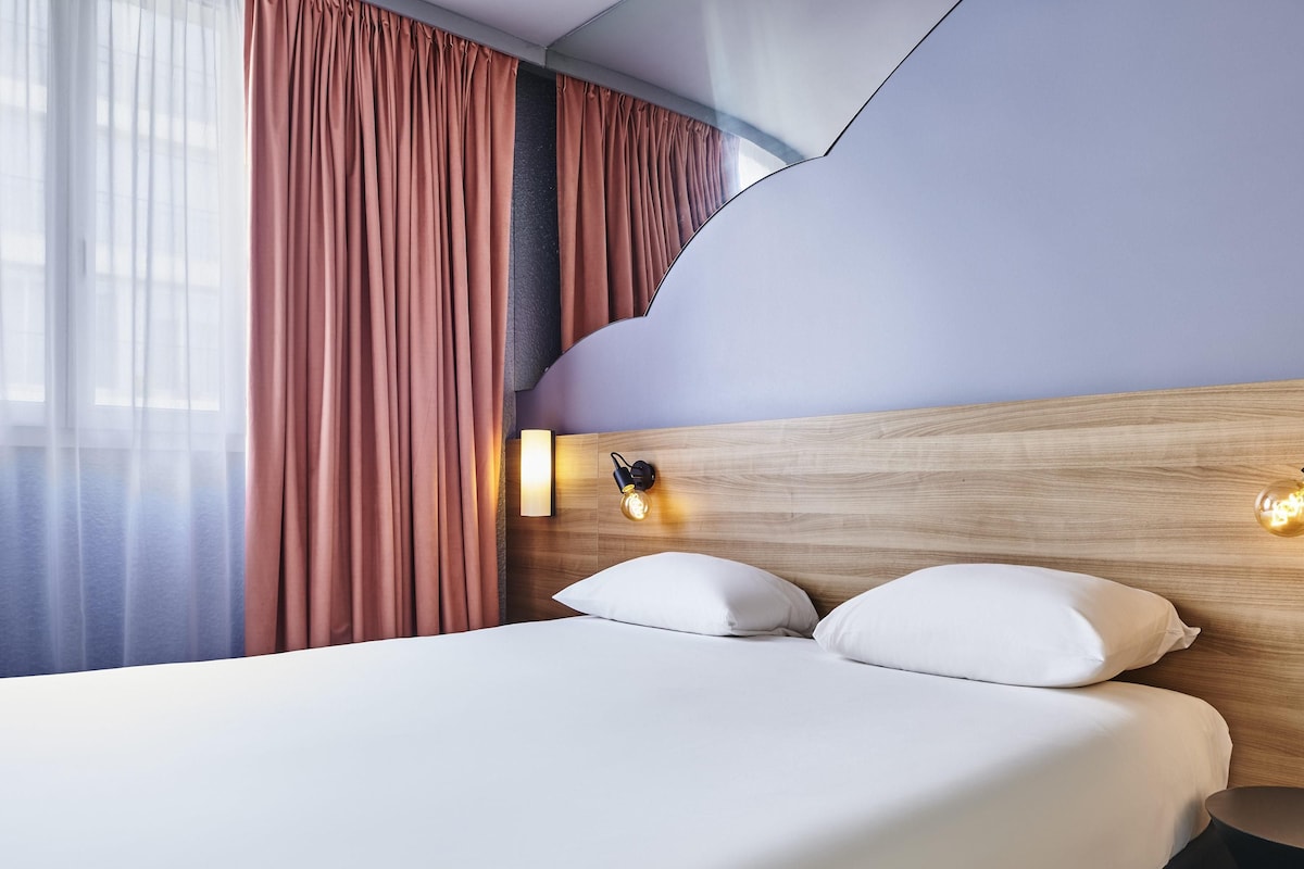 Habitación de hotel con cama, cortinas, una lámpara fijada a la pared y cabecero de madera.