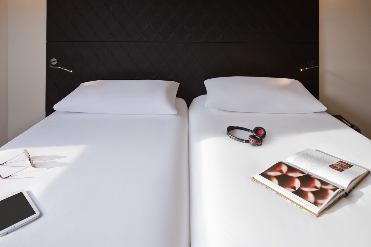 A double bed with white linens, a pair of glasses, a smartphone, and a magazine on the left side.
