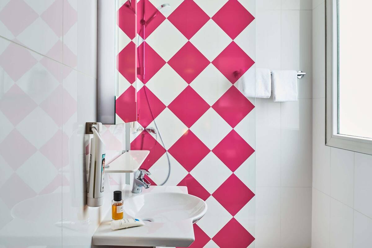 Une salle de bain avec un lavabo blanc, un miroir et une douche dont le mur est décoré d’un motif losangique rose et blanc.