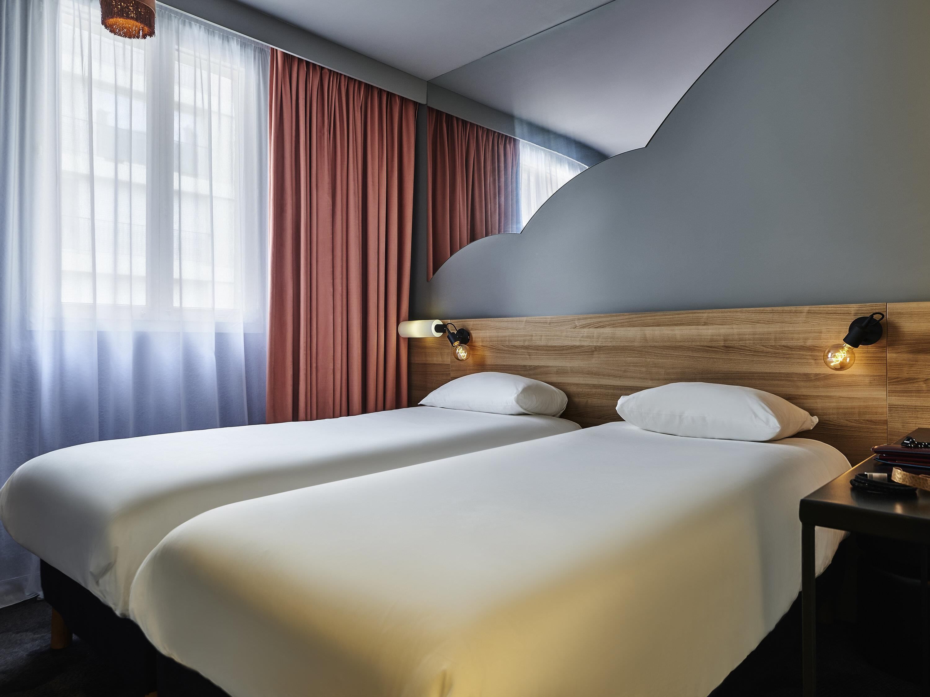 Hotellihuone, jossa on kaksi sänkyä, puinen pääty, yöpöytä ja ikkuna, jossa on verhot.