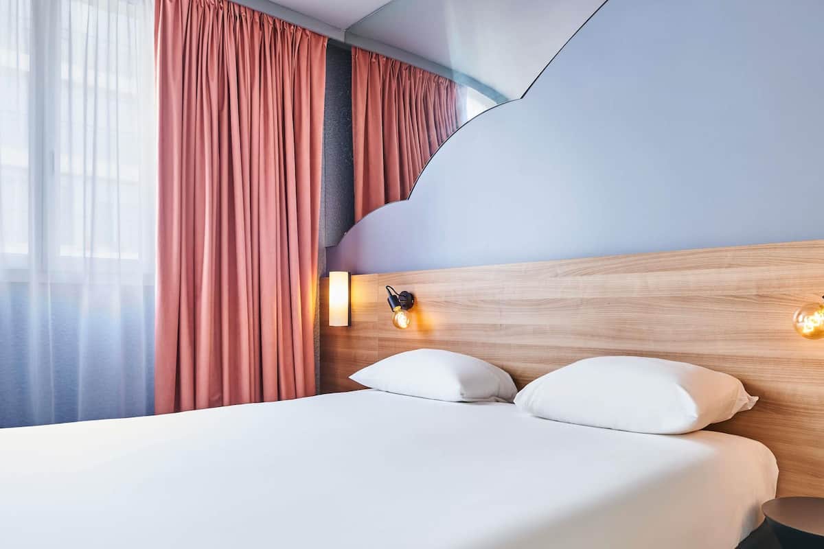 Een moderne hotelkamer met een houten hoofdeinde, een groot bed met witte lakens, en een nachtkastje met een lamp.