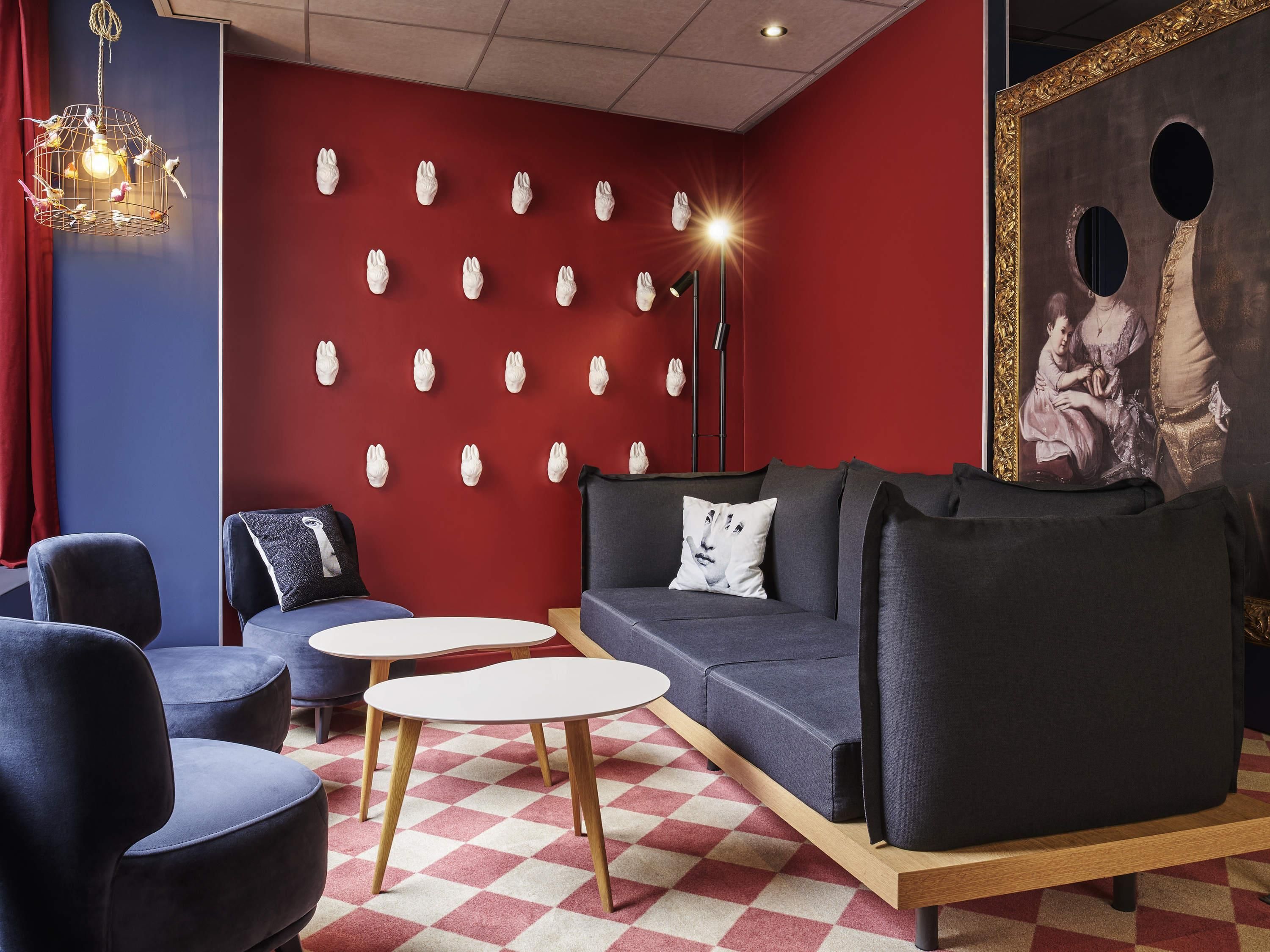 A modern lounge with a red wall, multiple seating options including a sofa and chairs, a patterned floor, and a decorative light fixture.