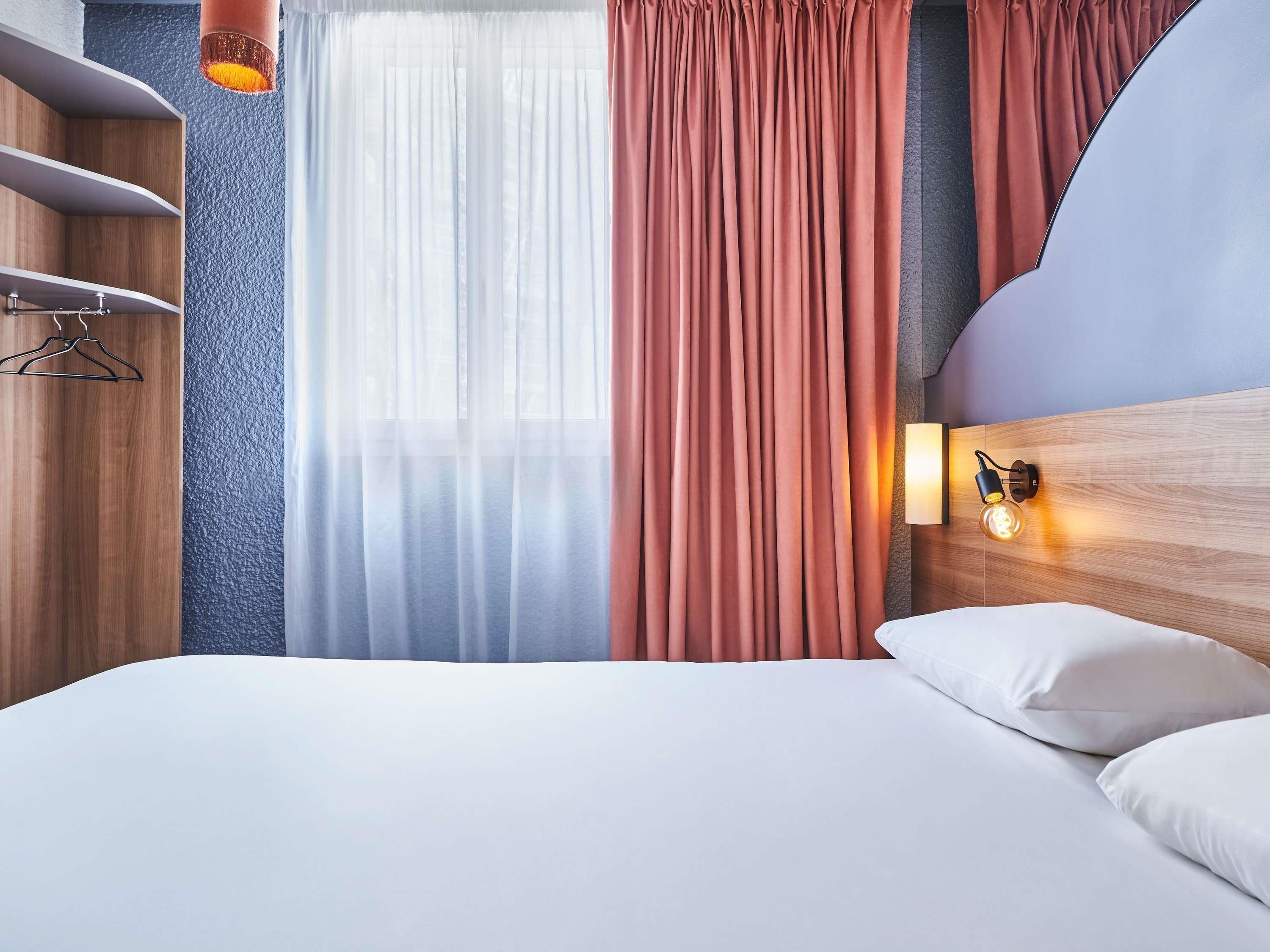 Une chambre d’hôtel avec un lit, une lampe fixée au mur et une fenêtre avec des rideaux légers.