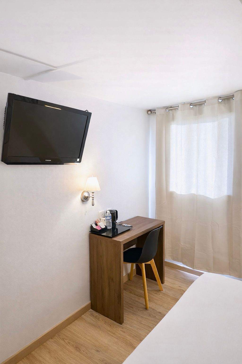 Una habitación de hotel con cama, escritorio con silla, televisor montado en la pared, lámpara y ventana con cortinas.