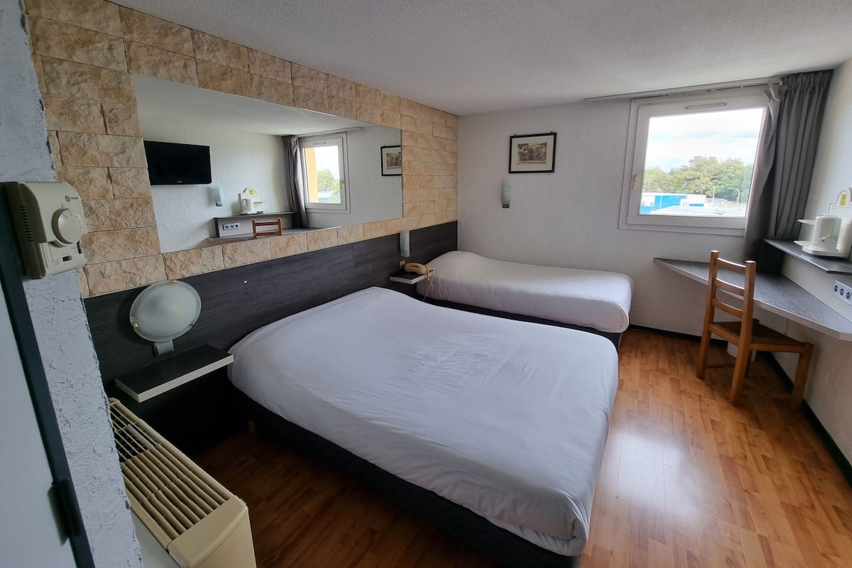 Ein Hotelzimmer mit zwei Betten, einem Schreibtisch, einem Stuhl, einem Heizkörper, einem Spiegel und einem Fenster mit Vorhängen.