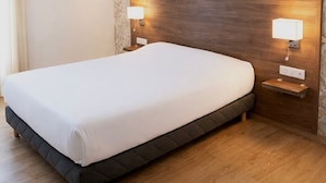 Habitación de hotel con una cama grande, cabecera de madera, dos lámparas de noche, un papel tapiz con motivos florales y una rejilla de ventilación en el techo.