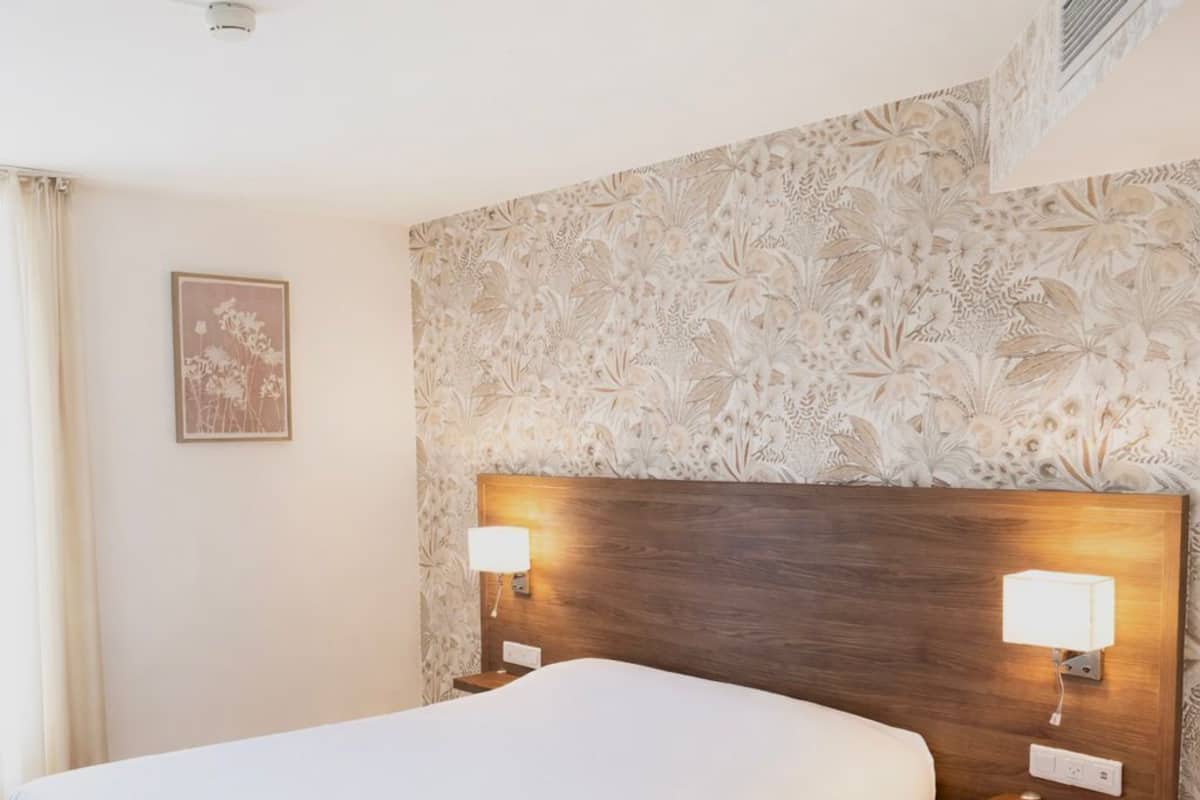 Ein Hotelzimmer mit einem großen Bett, einem Holzkopfstück, zwei Nachttischlampen, einer Wand mit Blumenmuster und einem Deckenlüfter.