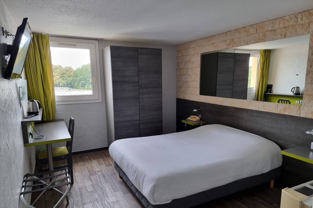 A modern hotel room with a bed, a desk, a chair, a mirror, and a window with curtains.