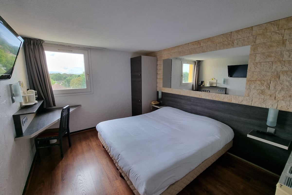 A hotel room with a bed, a desk, a chair, a window with curtains, a mounted TV, and a stone-textured wall.