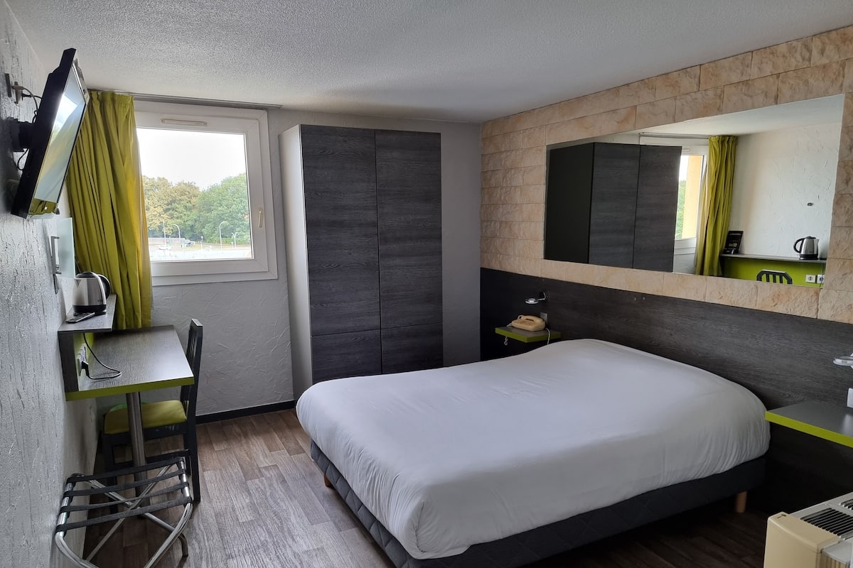 Ein modernes Hotelzimmer mit einem großen Bett, einem Schreibtisch mit Stuhl, einem kleinen Tisch, einem Spiegel und einem Fenster mit Vorhängen.