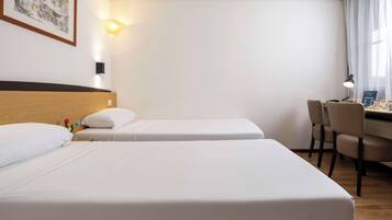 A hotel room with two single beds, a wooden headboard, a desk with a chair, and a wall-mounted lamp.