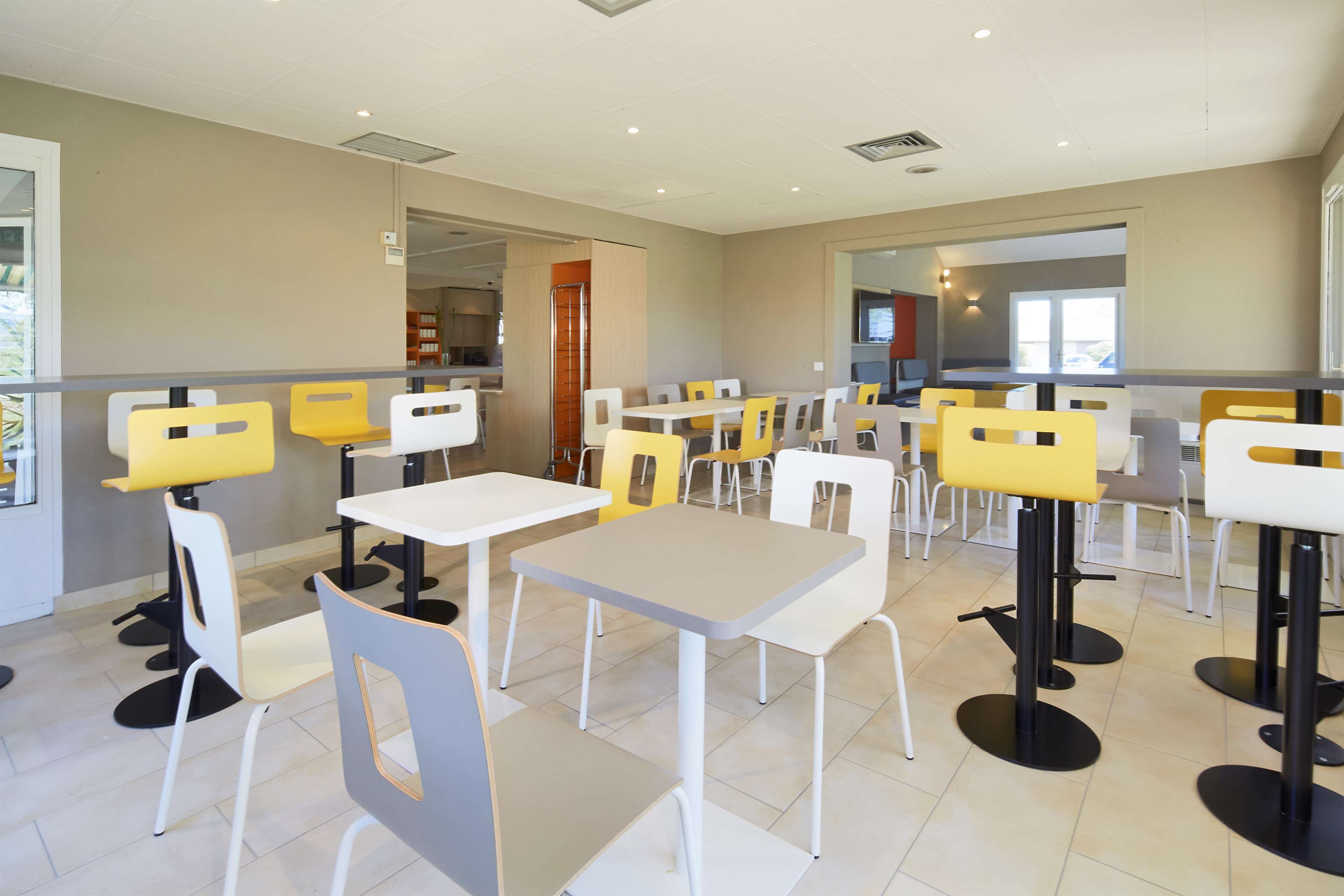 A modern dining area with white and yellow chairs, tables, and a kitchen in the background.