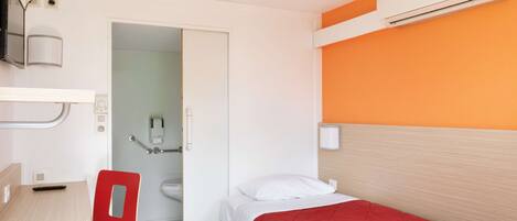 Habitación de hotel pequeña con una cama individual, un escritorio, una silla y un baño visible a través de una puerta abierta.
