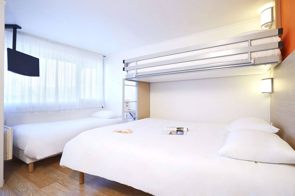 A hotel room with two beds, a flat-screen TV, and a window with sheer curtains.