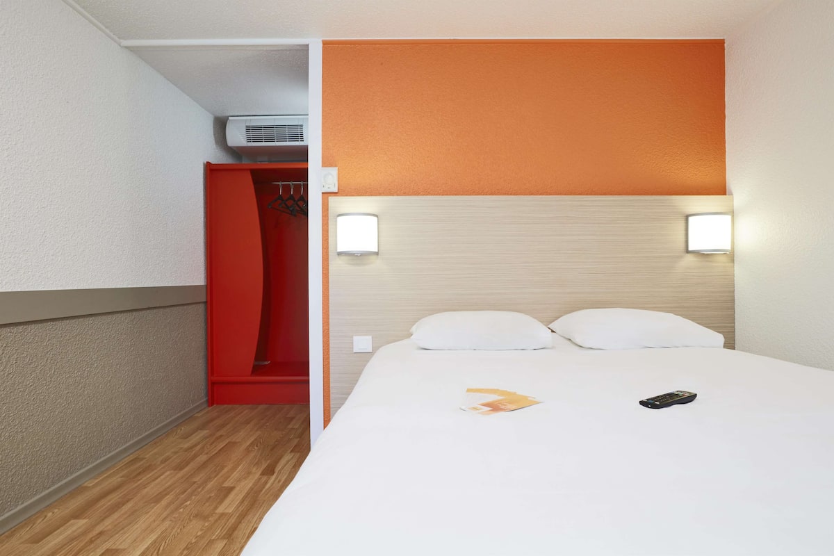 A hotel room with a white bed, two white pillows, a remote control, and a magazine on the bed. There is a red closet with a hanging rack and a white wall-mounted light.