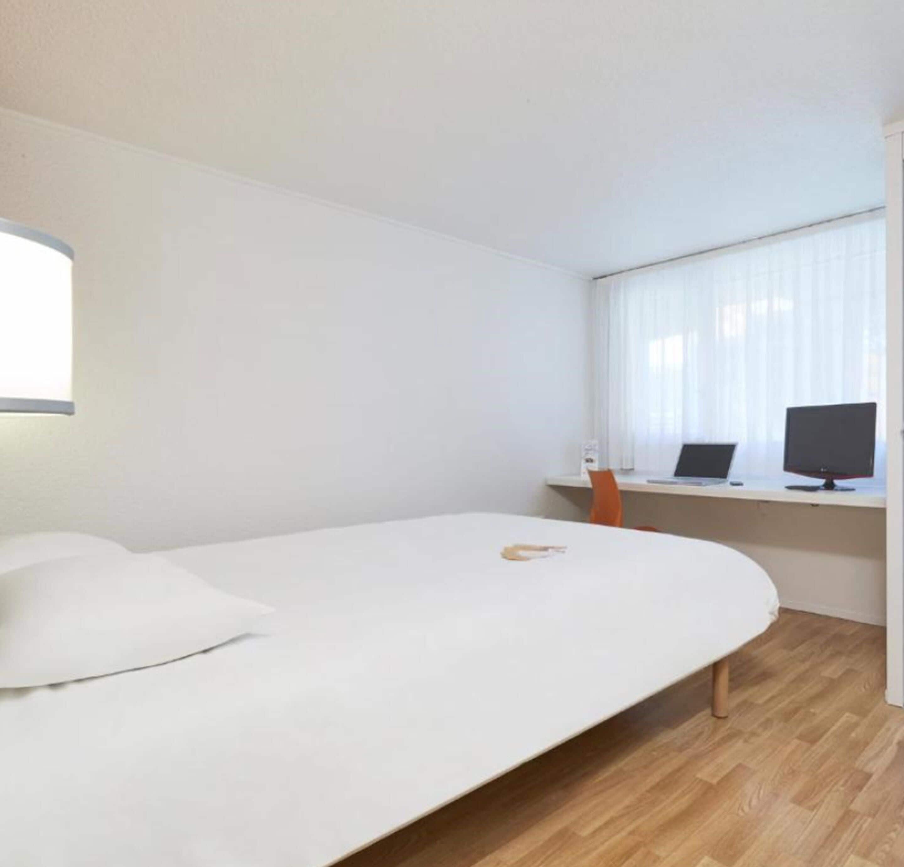 Et minimalistisk hotelværelse med en enkelt seng, et skrivebord med en computer og et vindue med let gardin.