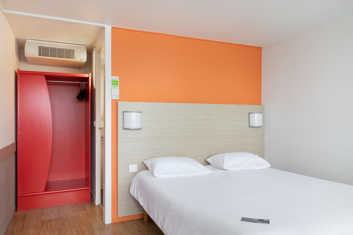 A hotel room with a bed, a red wardrobe, and a wall-mounted air conditioner.