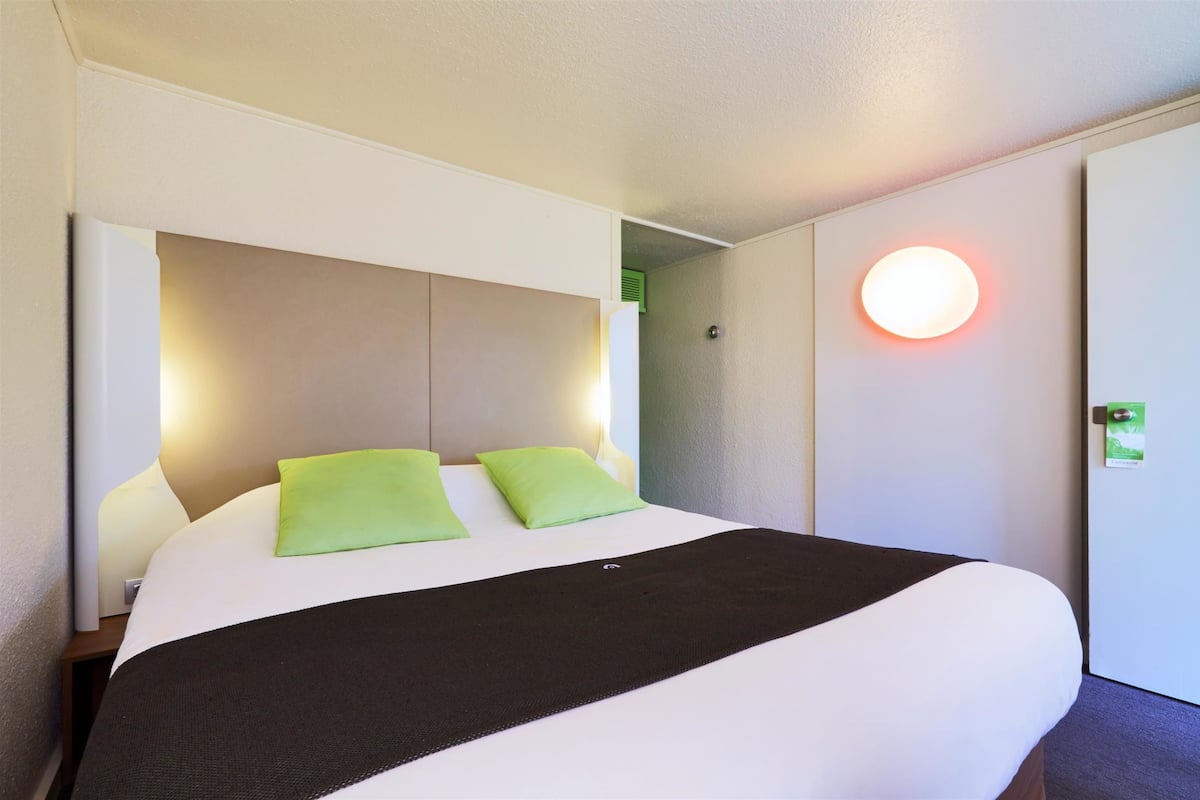 Una habitación de hotel con una cama grande, dos almohadas verdes, una manta de color negro y una lámpara de pared redonda.