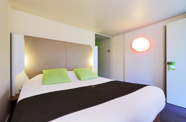 Una habitación de hotel con una cama grande, dos almohadas verdes, una manta de color negro y una lámpara de pared redonda.