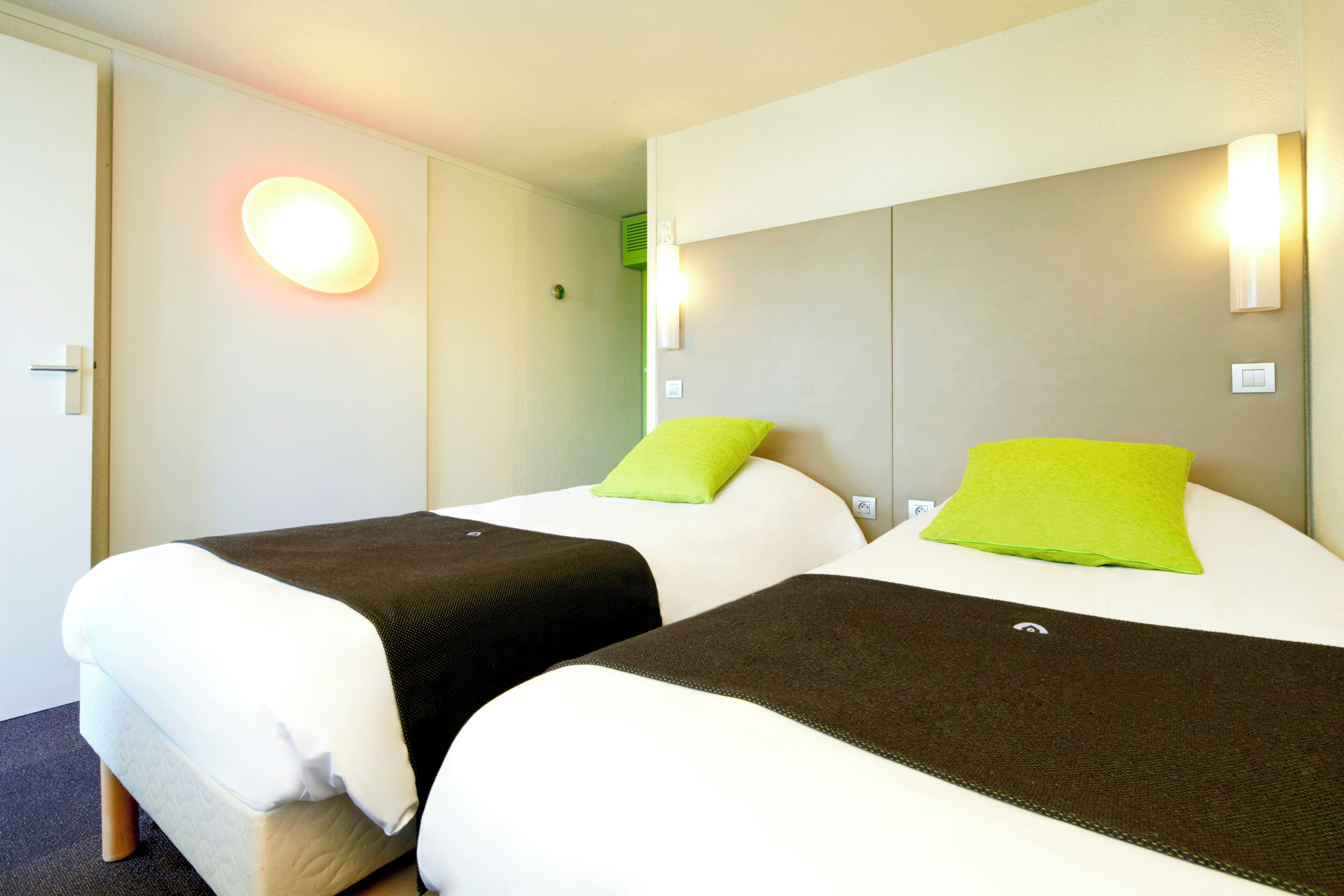 Dos camas con ropa de cama blanca y marrón, almohadas verdes y una lámpara de pared.