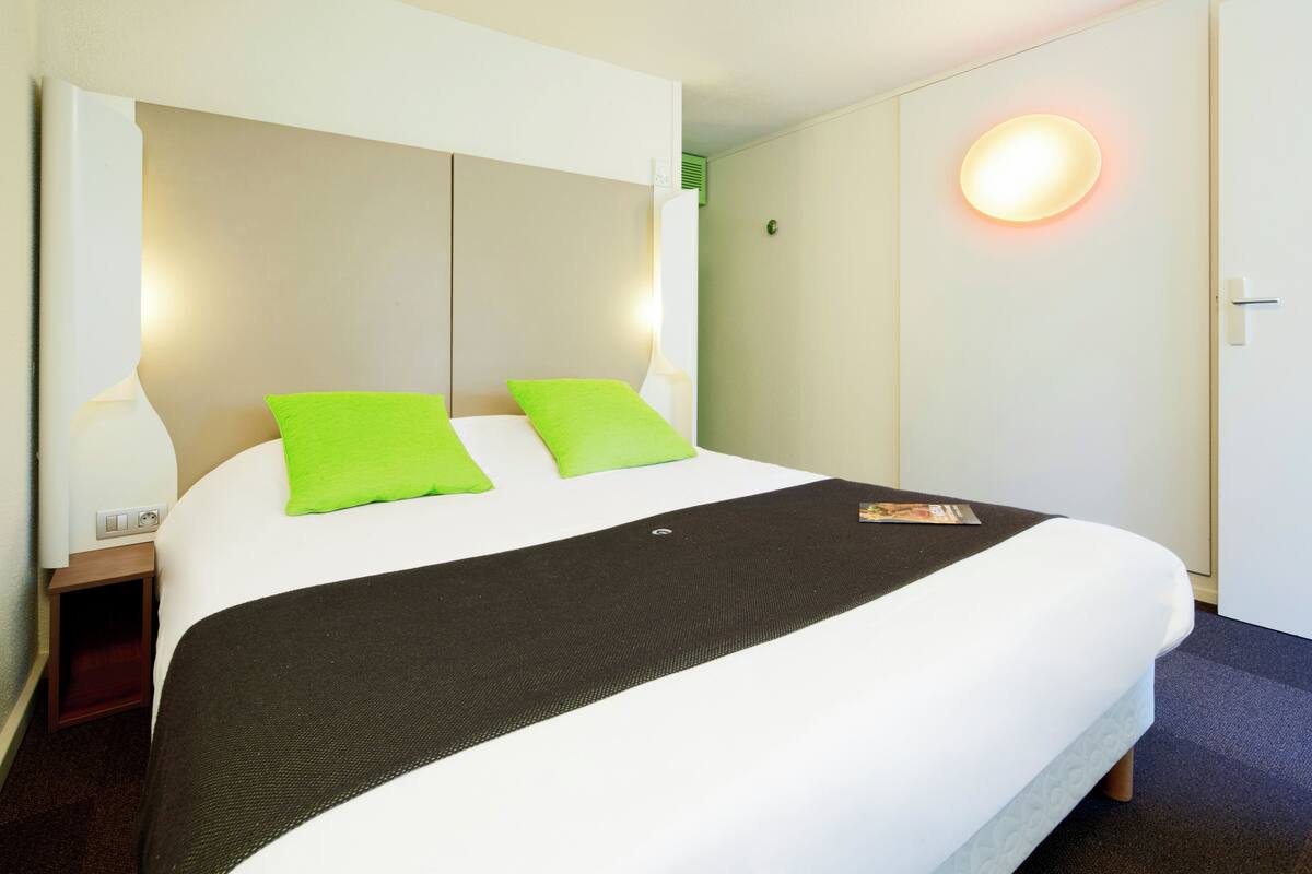 Habitación de hotel con una cama grande, dos almohadas verdes, una colcha negra y una lámpara de pared redonda.