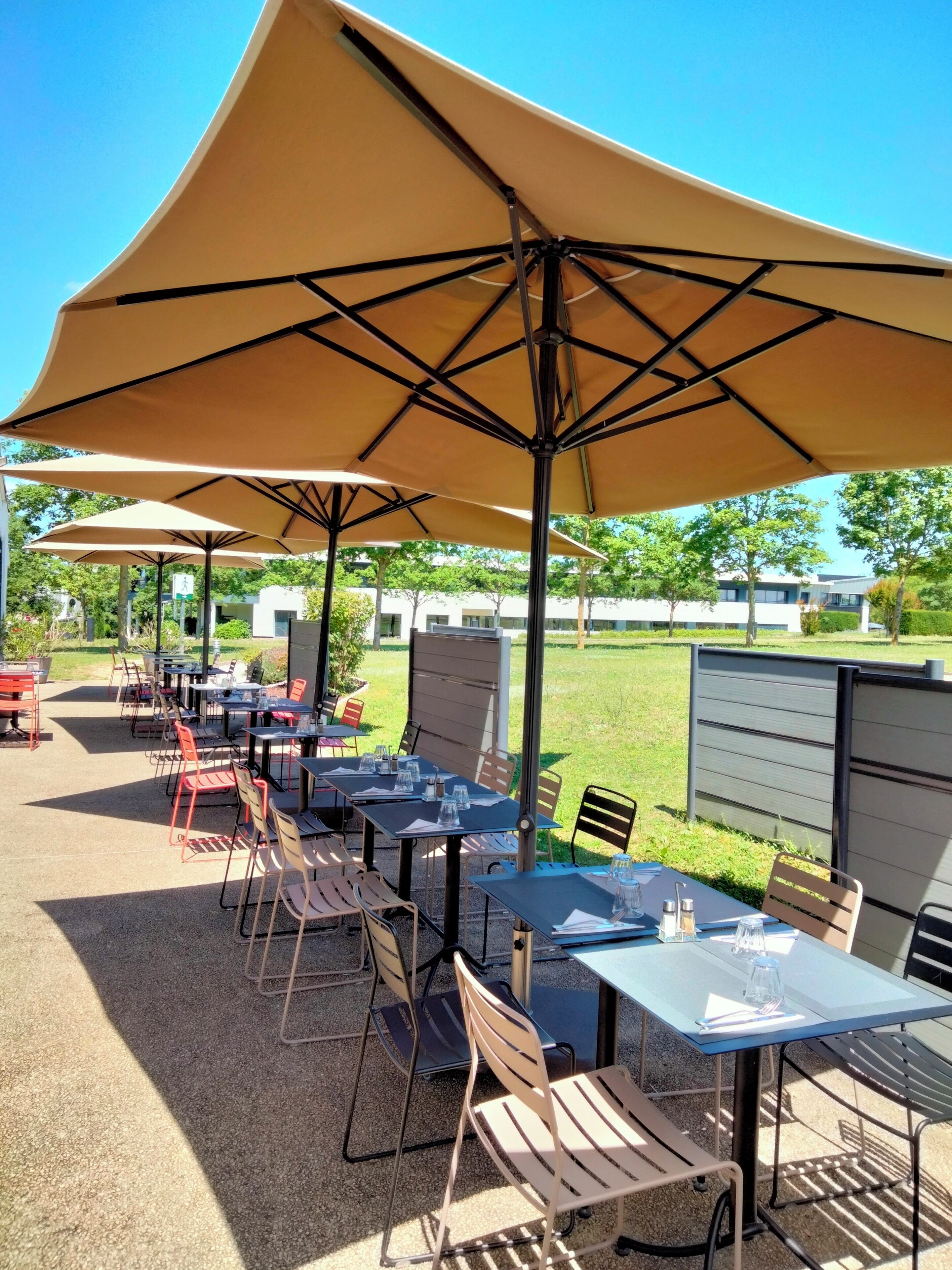 An outdoor patio area with tables and chairs under large umbrellas, surrounded by greenery and buildings in the background.