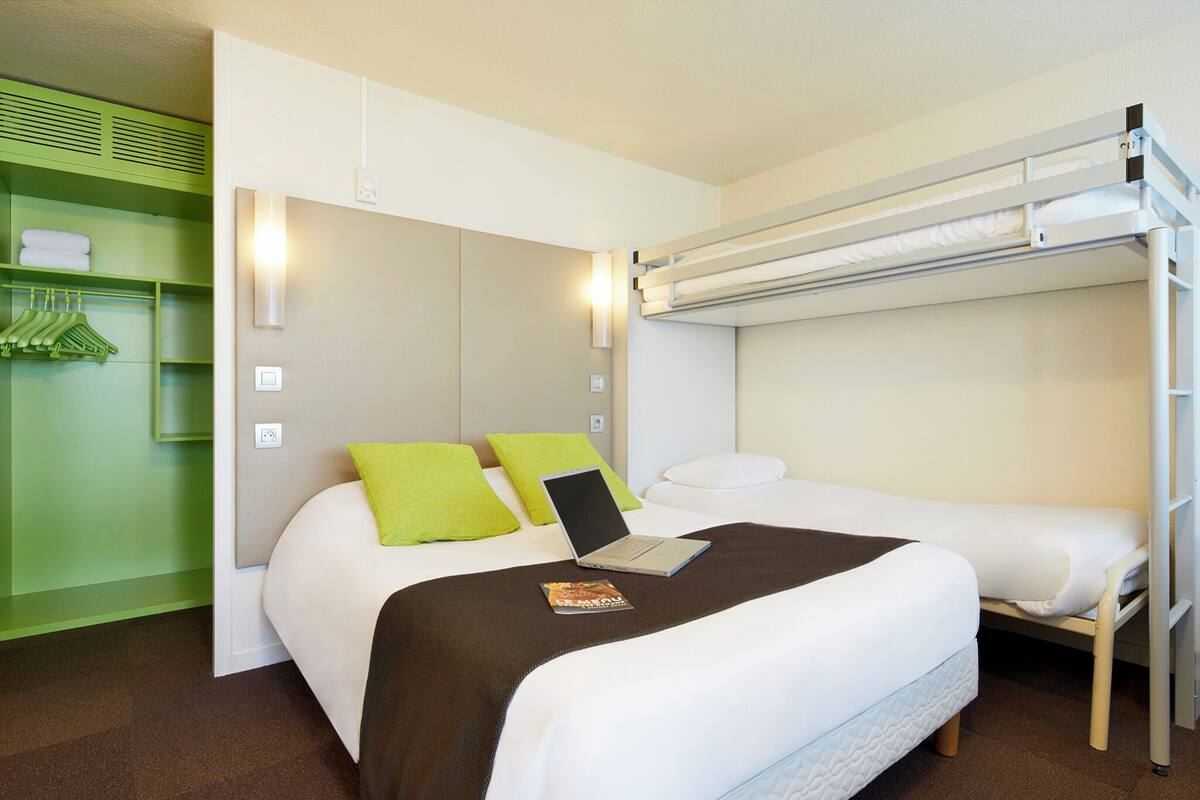 Habitación de hotel pequeña con una cama nido, una laptop sobre la cama inferior y un armario verde.