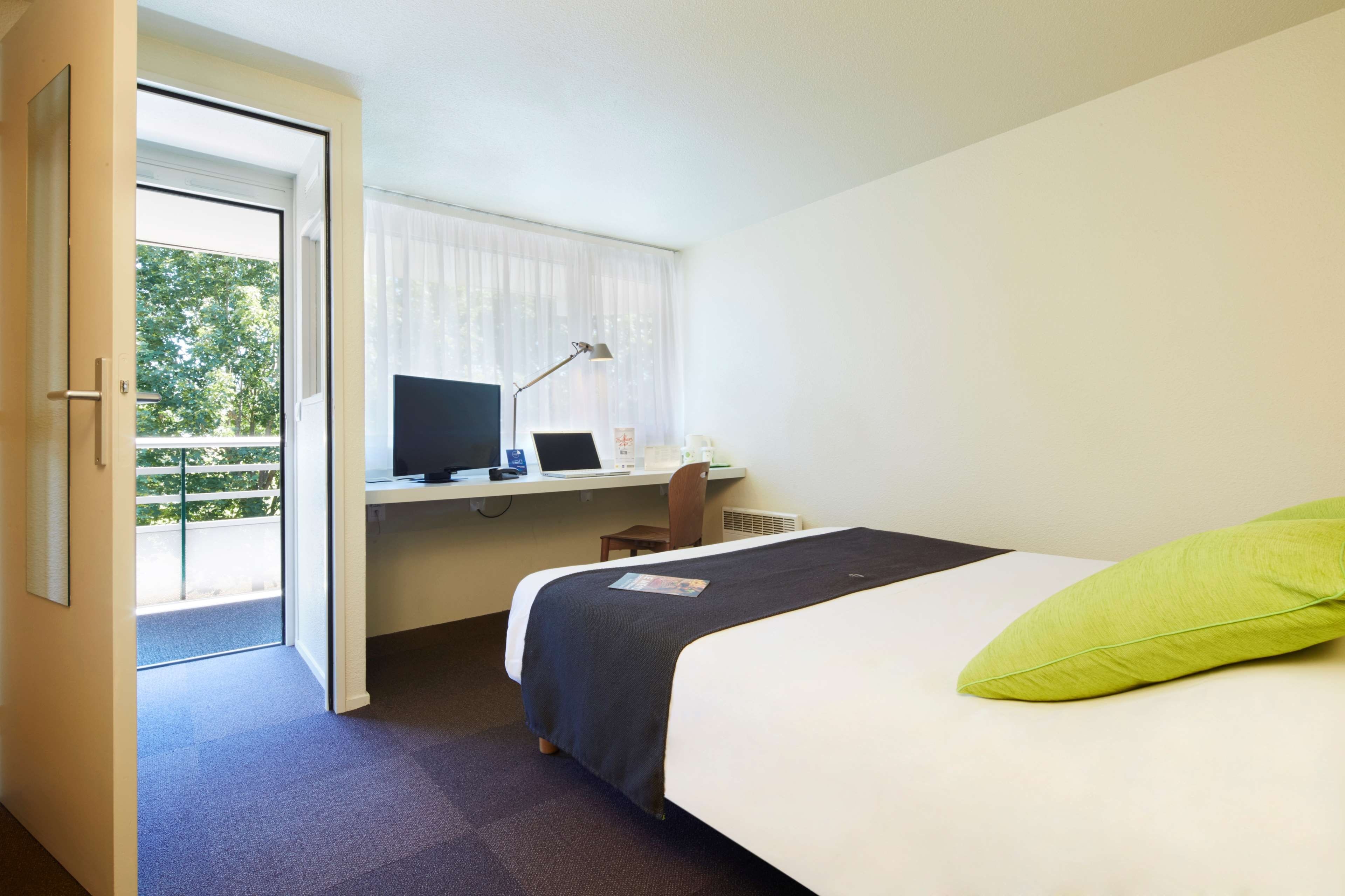 A hotel room with a bed, a desk with a computer, a chair, and a window with curtains.