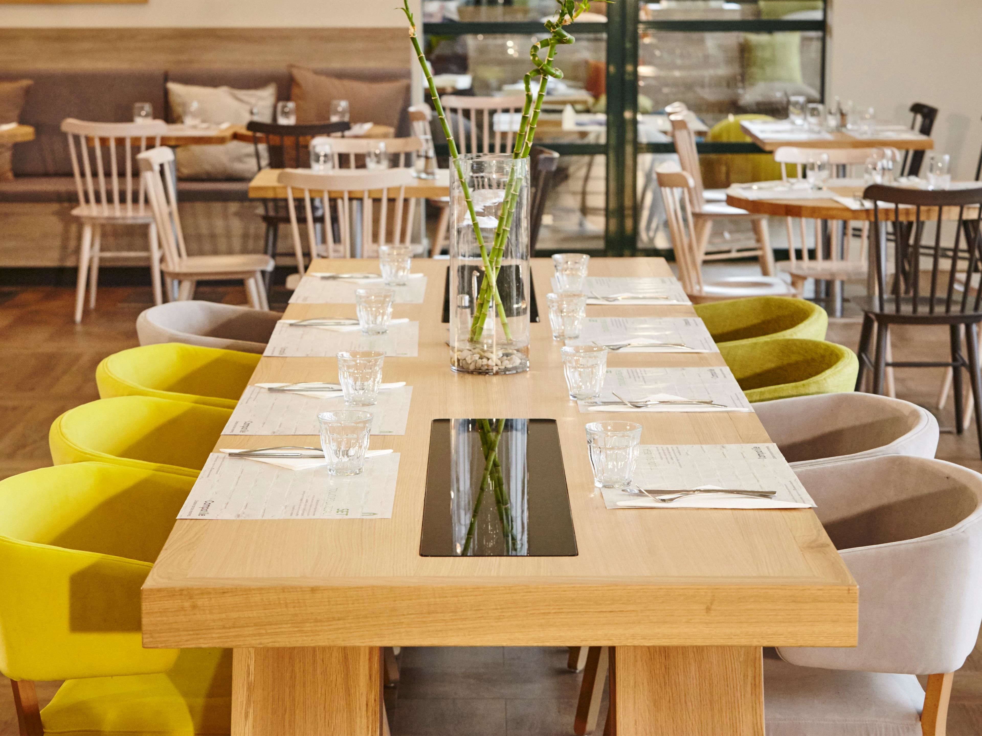Una larga mesa de comedor con múltiples sillas, cristalería transparente y un florero con plantas verdes.