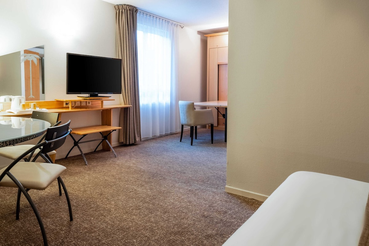 Une chambre d’hôtel équipée d’un téléviseur à écran plat, d’un bureau avec une chaise, d’une table à manger avec des chaises et d’un grand miroir.
