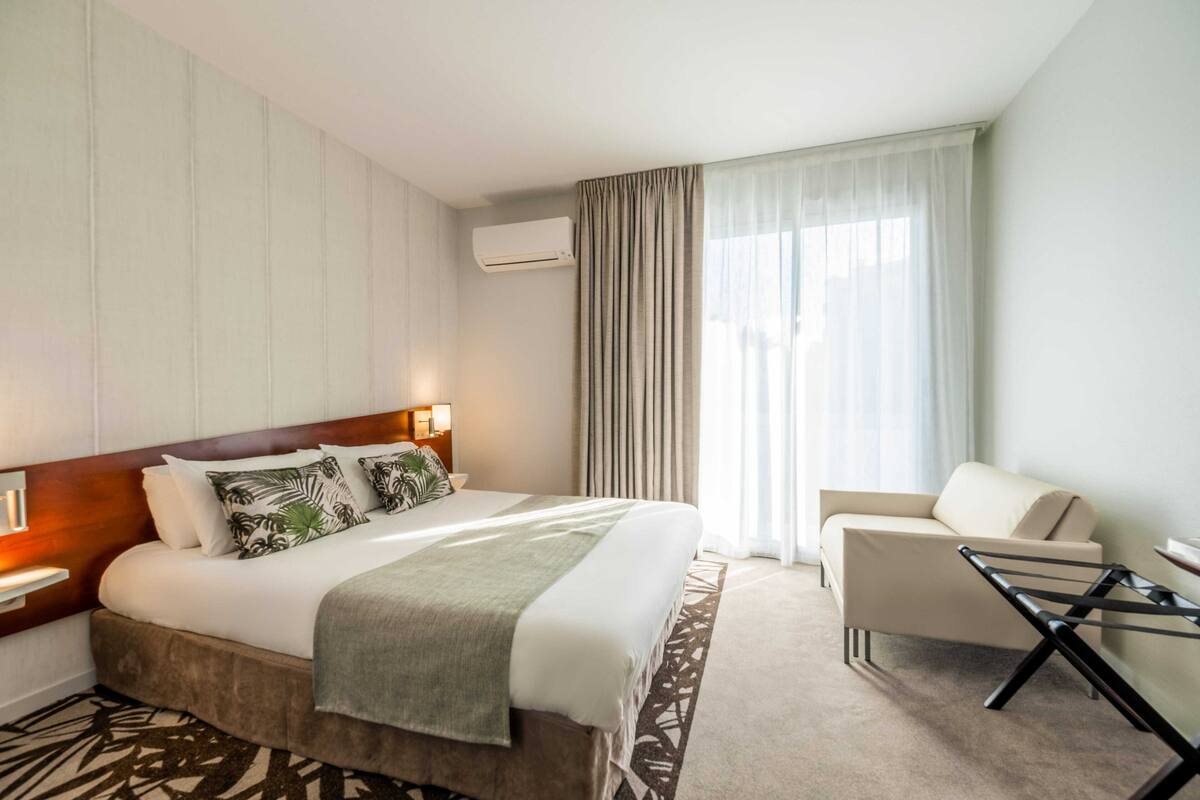 Une chambre d’hôtel avec un lit, une chaise, une petite table, un appareil de climatisation et une fenêtre avec des rideaux.