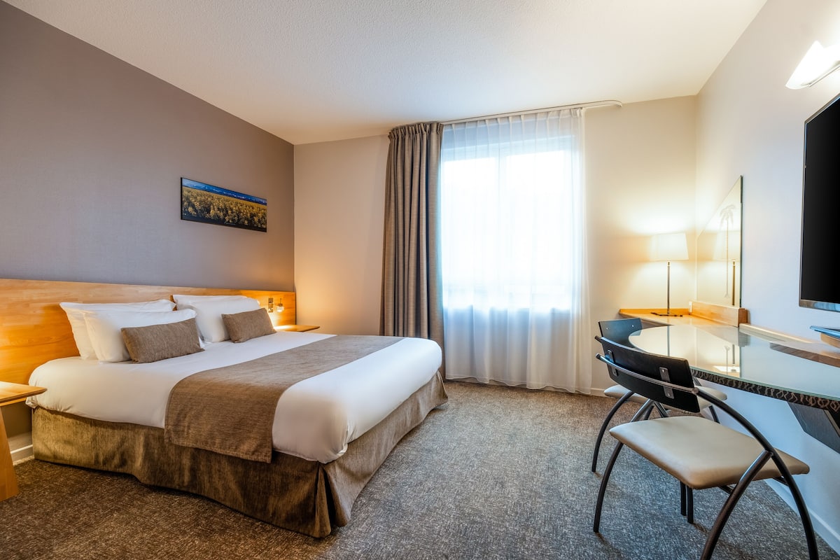 Une chambre d’hôtel avec un grand lit, un bureau avec une chaise, une télévision et une fenêtre avec des rideaux.