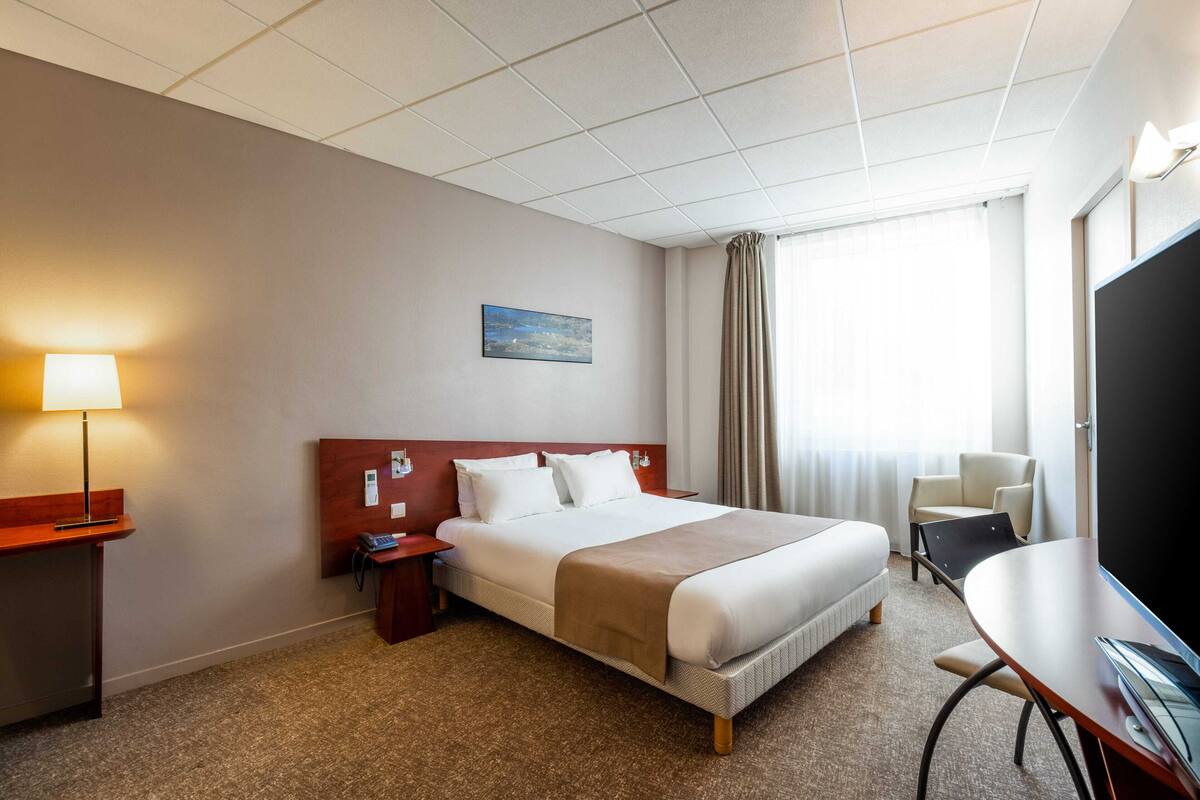Une chambre d’hôtel avec un grand lit, un bureau avec une chaise, une lampe, une télévision et un tableau au mur.