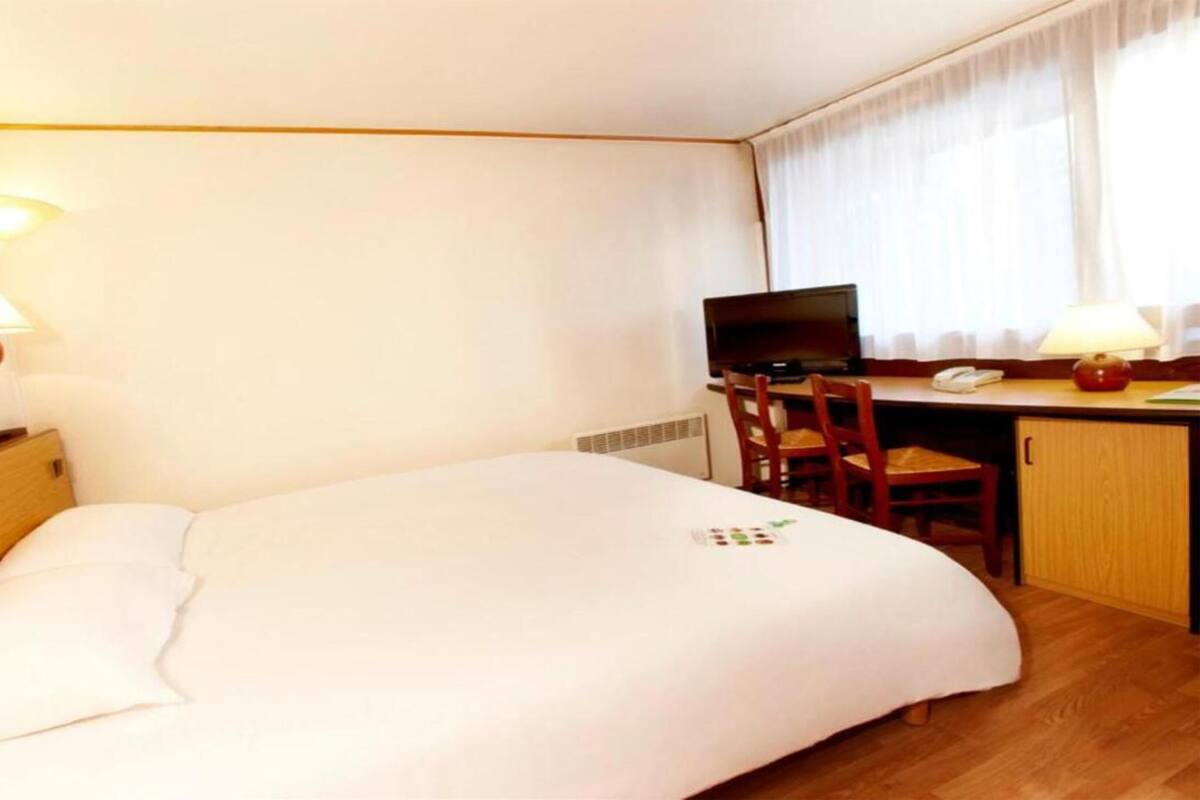Et hotelværelse med seng, skrivebord, stol, fjernsyn og et vindue med gardiner.