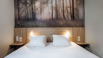 Een hotelkamer met een groot schilderij met bosmotief boven het bed, een houten hoofdbord, twee nachtkastjes met lampen en een witte sprei.