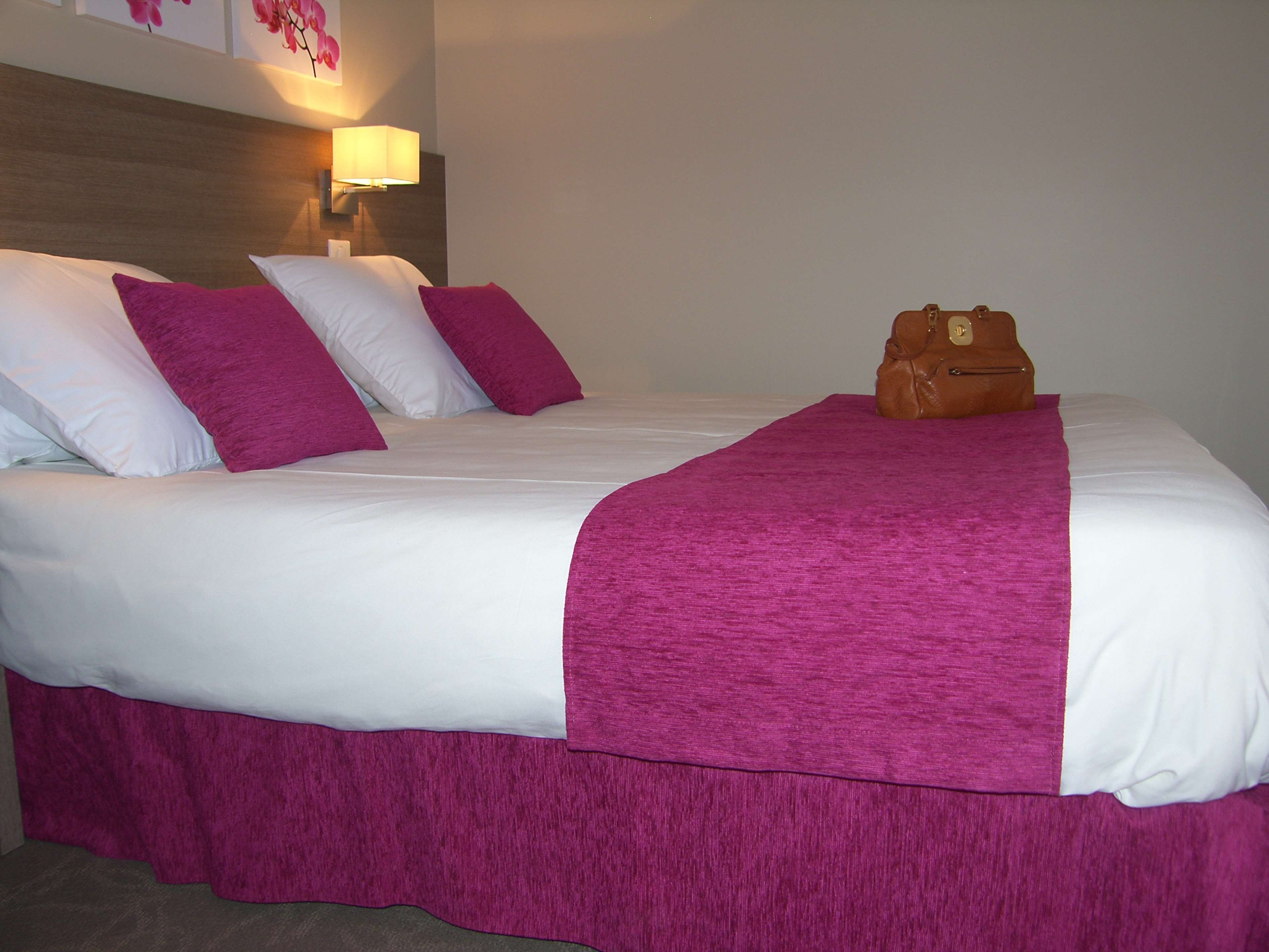 Ein ordentlich bezogenes Bett mit weißer und rosa Bettwäsche, eine braune Tasche auf dem Bett und eine Wandlampe.