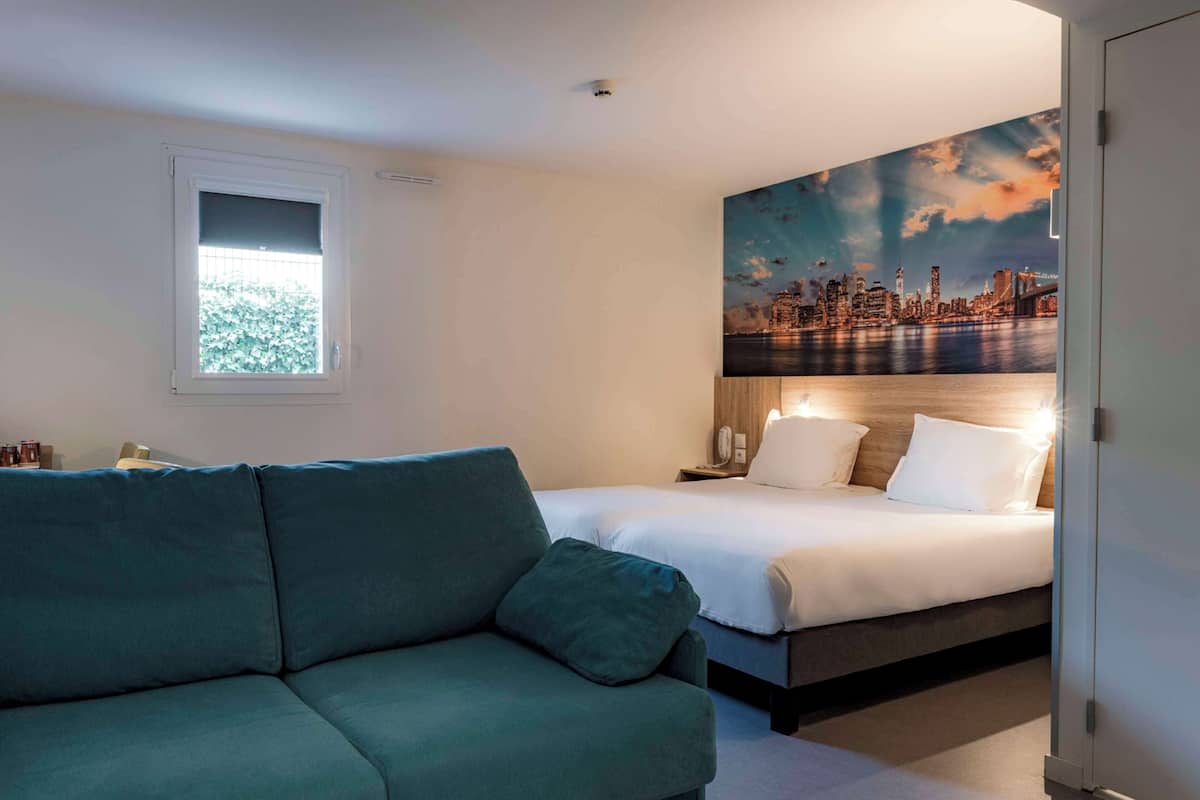 A bedroom with a bed, a green sofa, a cityscape mural, and a window.