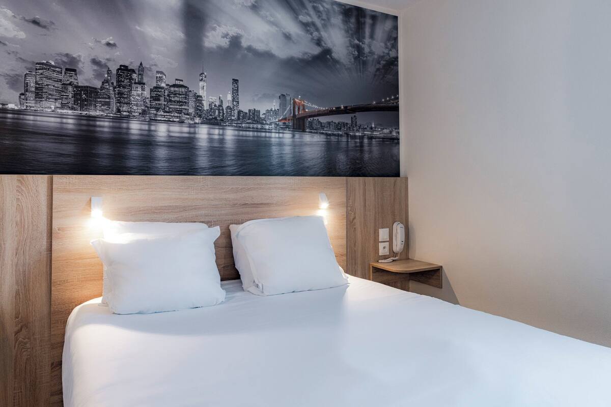A hotel room with a large bed, wooden headboard, white bedding, and a cityscape view on the wall.