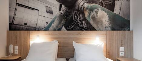 A hotel room with a large mural of an aircraft engine, two beds with white linens, wooden headboard, bedside tables, and wall sockets.