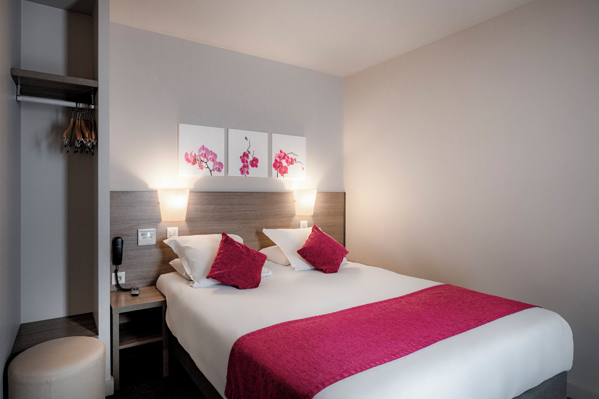 A hotel room with a bed, a nightstand, a lamp, a phone, and a wall-mounted shelf with hanging items.