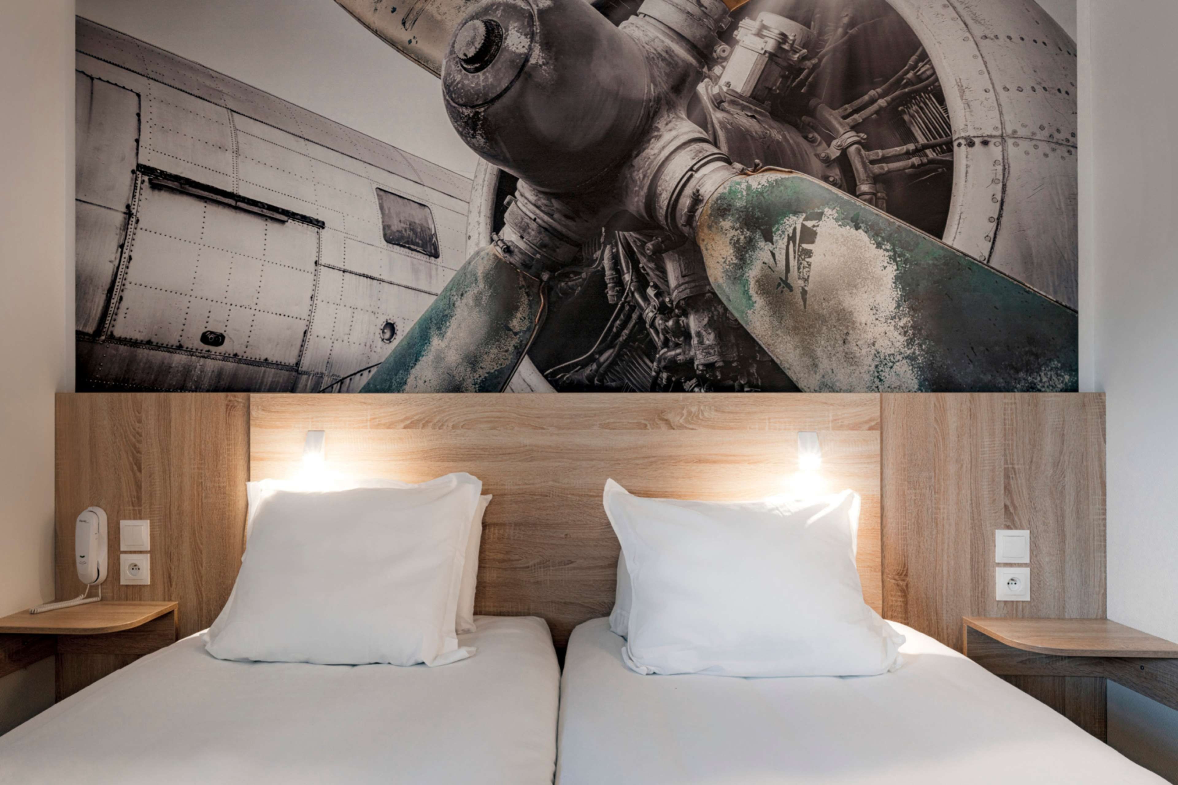 Hotelkamer met een groot muurschildering van industriële machines, een houten hoofdbord en twee nachtkastjes met lampen.