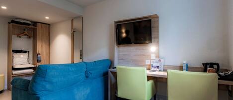 Habitación de hotel con sofá azul, dos sillones, un escritorio con televisor de pantalla plana, un armario de madera y una mesita con cafetera.