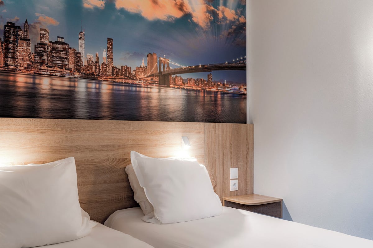 A hotel room with a bed, two pillows, a nightstand, and a wall-mounted light.