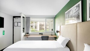 Une chambre d’hôtel comprenant un lit, un bureau, une chaise, une fenêtre avec des rideaux, une lampe fixée au mur et un tableau encadré.