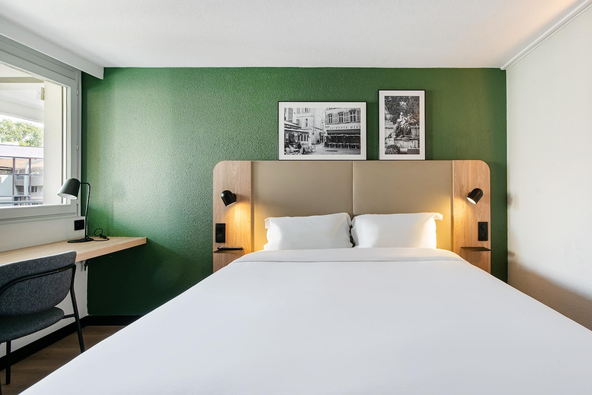 Hotelkamer met een groene accentmuur, een bed met witte lakens, twee bedlampen, een bureau met stoel, en twee ingelijste foto’s boven het bed.