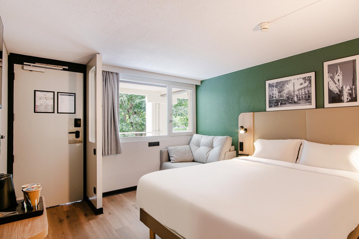 A hotel room with a large bed, a window with curtains, a green wall, and framed pictures.
