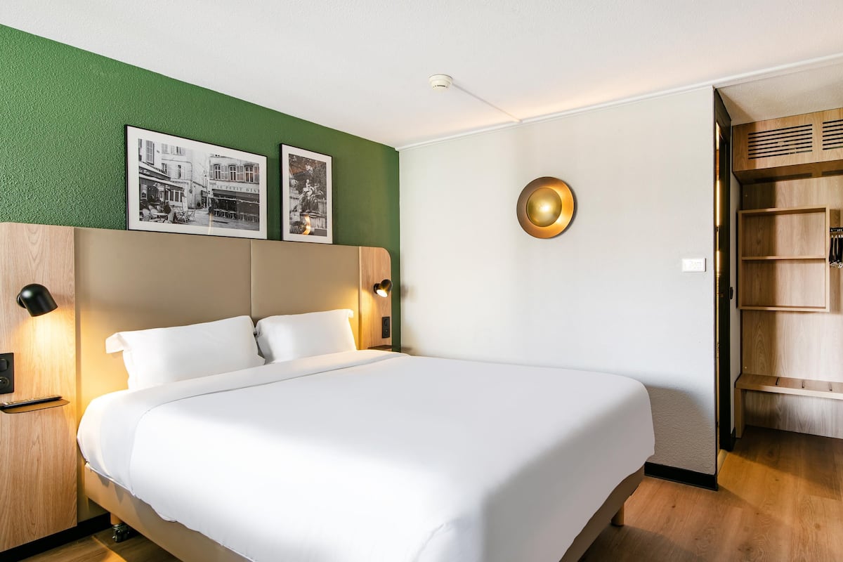 Een hotelkamer met een bed, een groene accentmuur, twee ingelijste foto's, een ronde wandklok en een houten garderobe.
