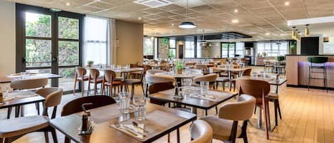 A spacious restaurant with multiple dining tables and chairs, large windows offering natural light, and a modern ceiling design.